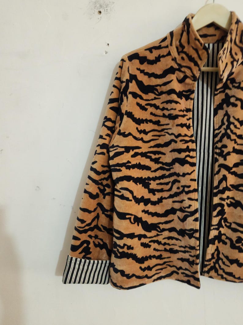 May include: A long-sleeved jacket featuring a tan and black tiger stripe pattern on the exterior. The interior lining and the folded cuffs display a contrasting black and white vertical pinstripe design. The jacket has a high collar and a front opening.