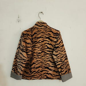 May include: A long-sleeved jacket featuring a tan and black tiger stripe pattern. The garment has a high collar and cuffs with a black and white vertical pinstripe design. The jacket is displayed on a hanger against a plain, light-colored wall.