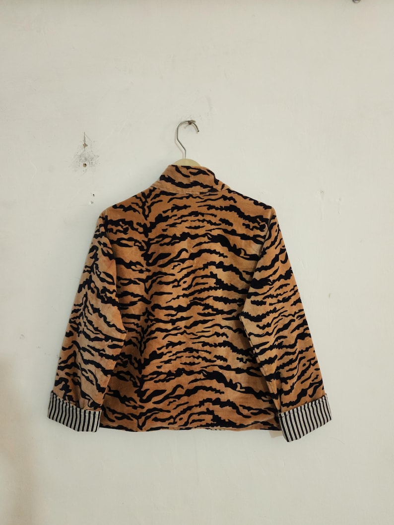 May include: A long-sleeved jacket featuring a tan and black tiger stripe pattern. The garment has a high collar and cuffs with a black and white vertical pinstripe design. The jacket is displayed on a hanger against a plain, light-colored wall.