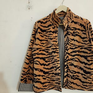 May include: A long-sleeve jacket featuring a tan and black tiger stripe pattern on the exterior. The garment has a high collar and a front opening that reveals a black and white vertical pinstripe lining, which is also visible on the sleeve cuffs.