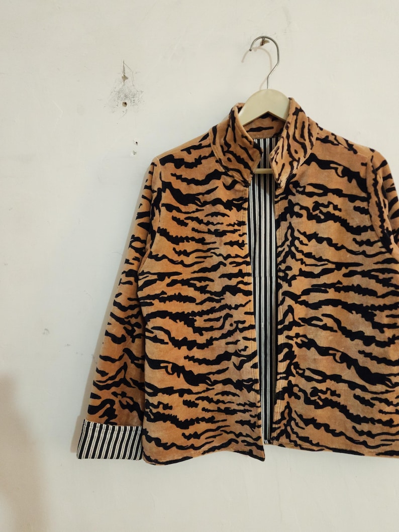 May include: A long-sleeve jacket featuring a tan and black tiger stripe pattern on the exterior. The garment has a high collar and a front opening that reveals a black and white vertical pinstripe lining, which is also visible on the sleeve cuffs.
