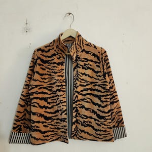 May include: A long-sleeved jacket featuring a tan and black tiger stripe pattern on the exterior. The jacket has a high collar, a front opening, and cuffs that reveal a black and white vertical pinstripe lining on the interior fabric.