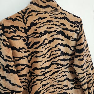 May include: A long-sleeved jacket featuring a tan and black tiger stripe pattern. The garment has a high collar and is constructed from a soft, textured fabric. The sleeves are finished with cuffs that display a black and white vertical stripe pattern.