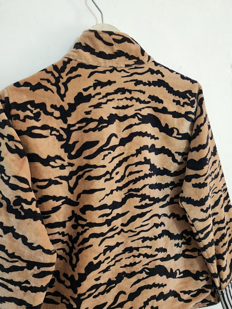 May include: A long-sleeved jacket featuring a tan and black tiger stripe pattern. The garment has a high collar and is constructed from a soft, textured fabric. The sleeves are finished with cuffs that display a black and white vertical stripe pattern.