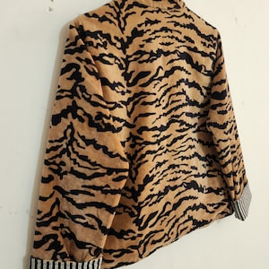 May include: A long-sleeved jacket featuring a tan and black tiger stripe pattern throughout. The garment has a high collar and cuffs that display a contrasting black and white vertical pinstripe design. The jacket is shown hanging on a hanger.