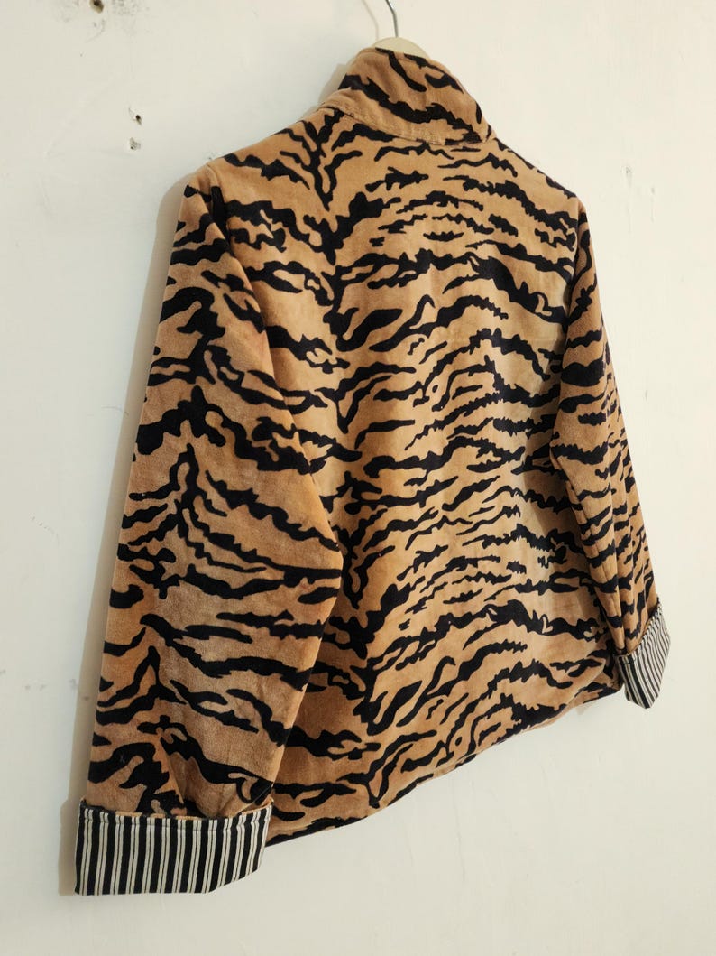 May include: A long-sleeved jacket featuring a tan and black tiger stripe pattern throughout. The garment has a high collar and cuffs that display a contrasting black and white vertical pinstripe design. The jacket is shown hanging on a hanger.
