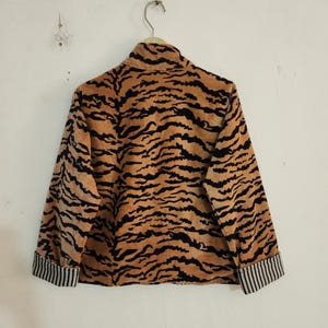 May include: A long-sleeved jacket featuring a tan and black tiger stripe pattern throughout. The garment has a high collar and cuffs with a black and white vertical pinstripe design. The jacket is displayed on a hanger against a plain wall.