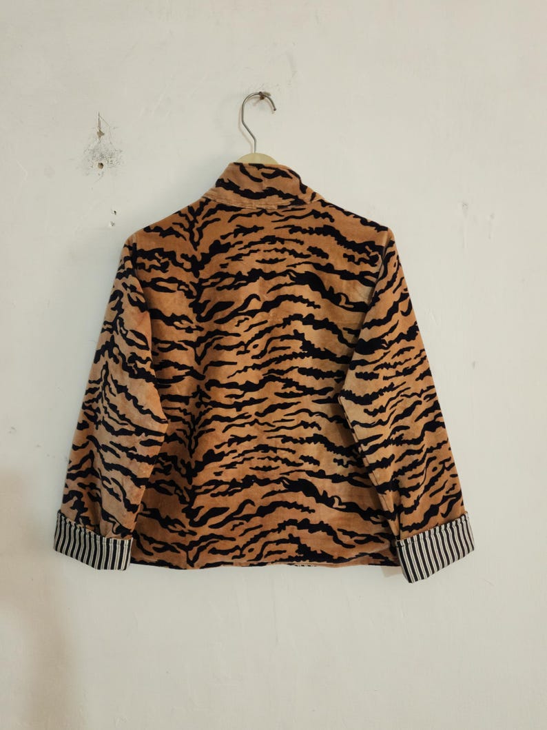 May include: A long-sleeved jacket featuring a tan and black tiger stripe pattern throughout. The garment has a high collar and cuffs with a black and white vertical pinstripe design. The jacket is displayed on a hanger against a plain wall.