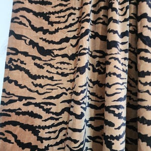 May include: A fabric with a tan base color featuring a repeating black tiger stripe pattern. The material is gathered in vertical folds, creating a textured appearance with light and shadow across the surface of the patterned textile.