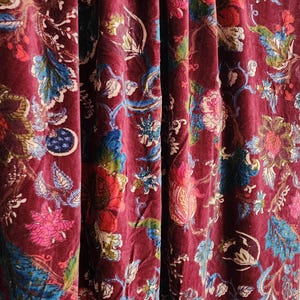 Op de afbeelding: Een diep bordeauxrode fluwelen stof met een dicht, veelkleurig bloemenpatroon. Het ontwerp bevat ingewikkelde botanische illustraties in tinten blauw, groen, roze en wit, met zachte plooien die diepte creëren op het textieloppervlak.