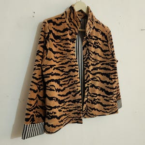May include: A jacket featuring a tan and black tiger stripe pattern on the exterior. The garment has a high collar, long sleeves with black and white striped cuffs, and an interior lining that displays a matching black and white vertical stripe pattern.