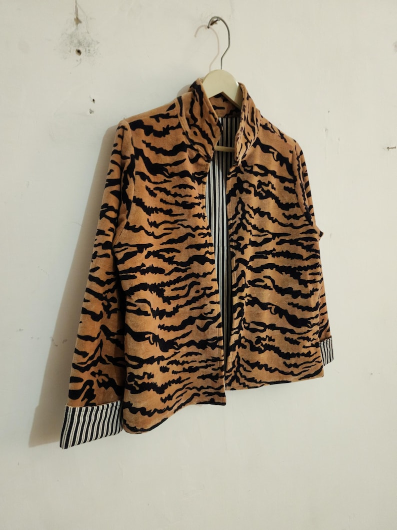 May include: A jacket featuring a tan and black tiger stripe pattern on the exterior. The garment has a high collar, long sleeves with black and white striped cuffs, and an interior lining that displays a matching black and white vertical stripe pattern.