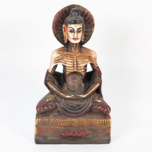 Könnte beinhalten: Eine sitzende Buddha-Statue mit Heiligenschein. Die Statue ist in Braun-, Rot- und Goldtönen bemalt. Der Buddha wird mit schlanker Statur und ruhigem Ausdruck dargestellt. Die Statue steht auf einem rechteckigen Sockel.