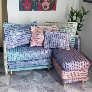 May include: A miniature chaise lounge sofa covered in rows of shimmering silver sequins. The piece includes a large light blue back cushion, a pink square cushion, and a small purple rectangular cushion, all featuring matching sequin detailing.