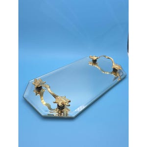 Puede incluir: Una bandeja octogonal de espejo con una superficie de vidrio reflectante y bordes biselados. La bandeja cuenta con dos asas de metal dorado ornamentadas, fijadas en los extremos opuestos, que presentan detalles decorativos intrincados.