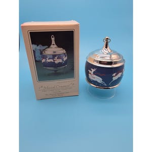 May include: A round musical ornament with a dark blue body featuring white reindeer with red ribbons, topped with a silver metallic cap and loop. It is displayed next to its original rectangular box labeled Musical Ornament First Christmas Together.