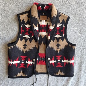 May include: A sleeveless vest featuring a geometric pattern in black, tan, red, and white. The garment has a shawl collar, black trim along the edges, and two silver-toned metal buttons at the front, each with a thin black leather cord attached.