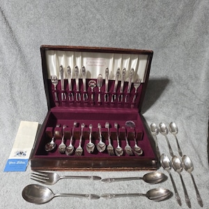 Könnte beinhalten: Ein versilbertes Besteckset in einem dunklen Holzkasten mit weißem Satin-Deckelfutter und einem tiefvioletten Samtboden. Das Set enthält verschiedene Gabeln und Löffel mit verzierten Griffen sowie eine kleine Informationsbroschüre mit der Aufschrift Your Silver.