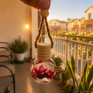 Op de afbeelding: Een bolvormig glazen flesje voor autoparfum, gevuld met heldere vloeistof en felroze bloemblaadjes. Het flesje heeft een geribbelde houten dop en hangt aan een dik, naturelkleurig touw van jute met een kleine houten kraal als accent.
