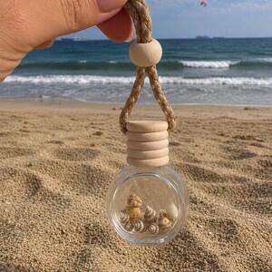 Peut inclure: Un petit flacon en verre transparent de forme ronde rempli de minuscules coquillages clairs. Le flacon est doté d'un bouchon en bois clair à rainures horizontales et est suspendu par un cordon tressé beige et doré avec une perle en bois.