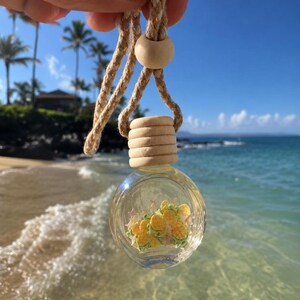 Könnte beinhalten: Eine kleine, runde, klare Glasflasche, gefüllt mit Flüssigkeit und gelben ananasförmigen Anhängern, mit einem hellen Holzdeckel und aufgehängt an einem dicken, geflochtenen beigen Seil mit einer passenden Holzperle als Akzent.