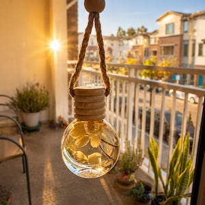 Op de afbeelding: Een kleine, bolvormige glazen fles gevuld met heldere vloeistof en witte bloemknoppen. De fles heeft een lichtgekleurde houten dop met een geribbeld ontwerp en hangt aan een dik, naturelkleurig touw van jute met een houten kraal aan de bovenkant.