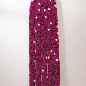 May include: A long, vertical garland of deep magenta flowers with scattered white blossoms, suspended from a hook. The floral arrangement is densely packed, creating a textured, eye-catching display. The background is a plain white wall.