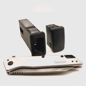 Puede incluir: Una navaja plegable Kershaw plateada con mango metálico y hoja visible, colocada frente a dos componentes rectangulares de plástico negro. La navaja presenta tornillos negros y una pieza de acento negra en el extremo del mango.