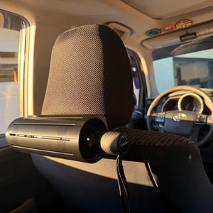以下が含まれることがあります： 車のヘッドレストの背面に水平に取り付けられた黒い円筒形の傘ホルダー。ホルダーの端にはテクスチャ加工が施されており、白い文字でTurnelleというブランド名が印刷されています。また、黒いリストストラップが下向きにぶら下がっています。