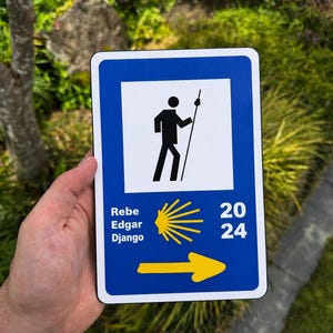 Puede incluir: Un letrero rectangular azul y blanco con el icono negro de un senderista con bastón. El letrero incluye los nombres Rebe, Edgar y Django, un gráfico de una vieira amarilla, el año 2024 y una flecha amarilla que apunta hacia la derecha.