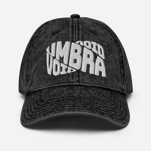 May include: A black, vintage-style baseball cap with the word "UMBRA VOID" in white, stylized lettering across the front. The cap has a curved brim and a button on top. The fabric has a worn, faded appearance.