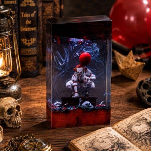 Può includere: Un blocco rettangolare in resina contenente una miniatura di clown accovacciata su una piattaforma scura, che tiene in mano un palloncino rosso. L'interno presenta effetti di fumo bianco vorticoso, con base e parte superiore color rosso.