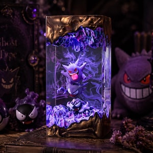 May include: A rectangular resin display piece featuring three stacked purple Gengar figures surrounded by swirling purple smoke effects, encased in a clear block with textured, metallic bronze-colored top and bottom edges.