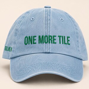 May include: A light blue denim baseball cap with a six-panel construction and a curved brim. The front features the embroidered green text ONE MORE TILE, while the side panel includes the smaller embroidered green text DEAL ME IN.