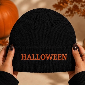 以下が含まれることがあります： 前面にオレンジ色のアルファベット大文字で「HALLOWEEN」と刺繍された、折り返し付きの黒いニット帽。リブ編みの生地で作られており、全体が単色の黒で統一されたデザインの帽子です。