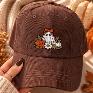 May include: A brown corduroy baseball cap featuring an embroidered design of a white ghost wearing an orange bow, surrounded by orange and white pumpkins, green leaves, and white daisy flowers centered on the front panel of the hat.