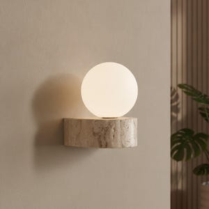 May include: A wall-mounted light fixture featuring a spherical white glass globe shade resting on a semi-circular base made of beige, porous natural stone with a textured, pitted surface that displays organic veining and earthy color variations.