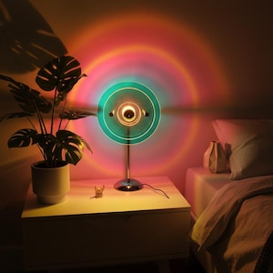 May include: A circular sunset projection lamp with a chrome base and stand, emitting a vibrant gradient of teal, pink, and orange light in concentric rings against the wall. The lamp features a clear glass lens and a thin, metallic support structure.
