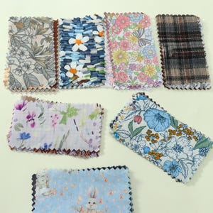 May include: A collection of seven rectangular fabric swatches with pinked edges. Patterns include grey botanical prints, blue and white daisies, colourful floral motifs, a dark plaid, a light floral watercolour, blue flowers, and a light blue rabbit print.