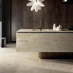 May include: A modern kitchen island with a light-colored stone countertop and a black faucet. The island has a brass base and is set against dark wood cabinetry. A white floral chandelier hangs above the island, illuminating the space. The floor is tiled in a matching light stone.