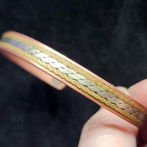 Op de afbeelding: Een armband met een centrale band van gevlochten zilverkleurig metaal, geflankeerd door twee dunne goudkleurige gedraaide draadaccenten, geplaatst in een glad koperkleurig metalen frame met een zichtbare ronde magneet aan het uiteinde.