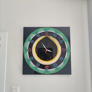 May include: A square wall clock featuring a black background with intricate string art patterns. The design includes concentric circles of yellow, white, and green thread, accented by small circular motifs in red and blue. White and red clock hands.