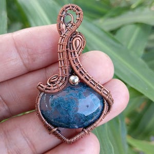 Può includere: Un ciondolo con una pietra di agata muschiata liscia e arrotondata con inclusioni blu-verde intenso, incastonata in una complessa cornice di filo di rame avvolto. Il design include una piccola perla metallica lucida nella struttura.