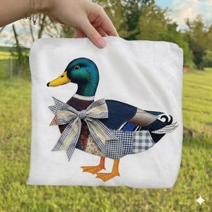 May include: A white fabric square featuring a mallard duck appliqué. The duck has a metallic green head, a yellow beak, and a body composed of various fabric patches including blue gingham and floral patterns, finished with a large fabric bow.