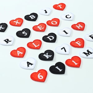 May include: A collection of heart-shaped embroidered patches in red, black, and white. Each patch features a single letter or number stitched in a contrasting color. The patches are scattered across the frame, displaying various characters including letters like A, B, C, D, E, F, H, I, J, K, L, M, R, X and numbers like 1, 5, 6, 7.