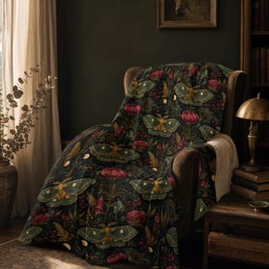 May include: A dark-colored throw blanket featuring a repeating pattern of green moths, deep pink flowers, and small golden moon phases. The blanket has a soft, light-colored sherpa-style lining visible on the edge, draped over a dark brown armchair.