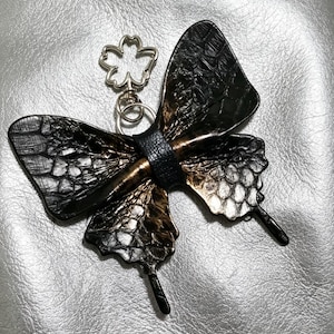 Puede incluir: Un llavero con forma de mariposa que presenta alas texturizadas en negro y bronce con un patrón de escamas. El centro está envuelto en una banda negra texturizada e incluye un cierre de metal plateado con forma de flor de cinco pétalos.