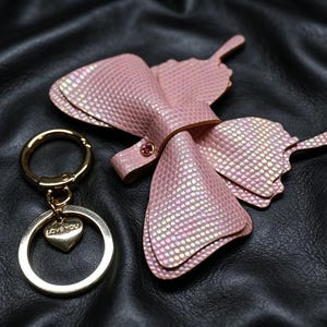 Puede incluir: Un accesorio con forma de mariposa rosa con un acabado texturizado e iridiscente y un pequeño remache de metal dorado, junto a un llavero de metal dorado con un dije en forma de corazón grabado con las palabras "LOVE YOU".