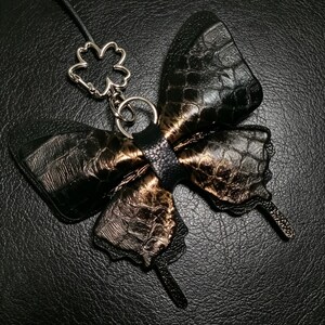 May include: A butterfly-shaped charm featuring a black and metallic bronze textured finish with a crocodile-like pattern. The charm is attached to a silver-toned metal clover-shaped clasp and a thin black cord, creating a decorative accessory.