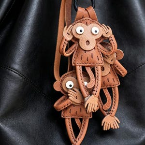 Peut inclure: Un porte-clés en cuir beige représentant trois singes dans les postures ne rien voir, ne rien entendre et ne rien dire. Chaque singe présente des détails cousus, des yeux circulaires blancs avec des pupilles noires et de longs membres.